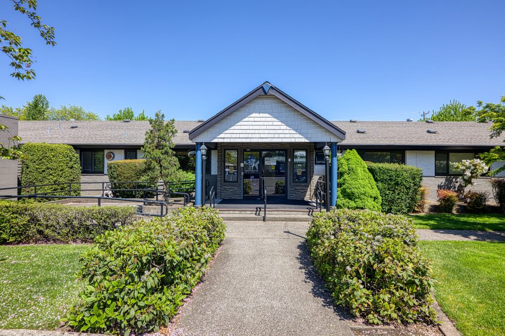 The front entrance of a building with a covered walkway, a stone facade, and a walkway surrounded by green bushes